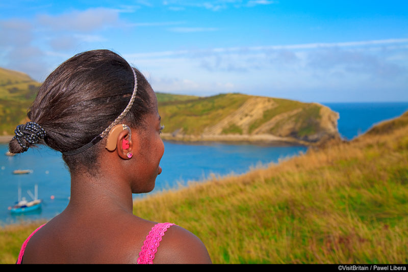 A woman admiring the view from the clifftop above Lulworth Cove on Dorset's Jurrassic coast. The woman is wearing a hearing aid and is profoundly deaf., Lulworth Cove, Dorset, England. Additional Credit: Tourism For All
