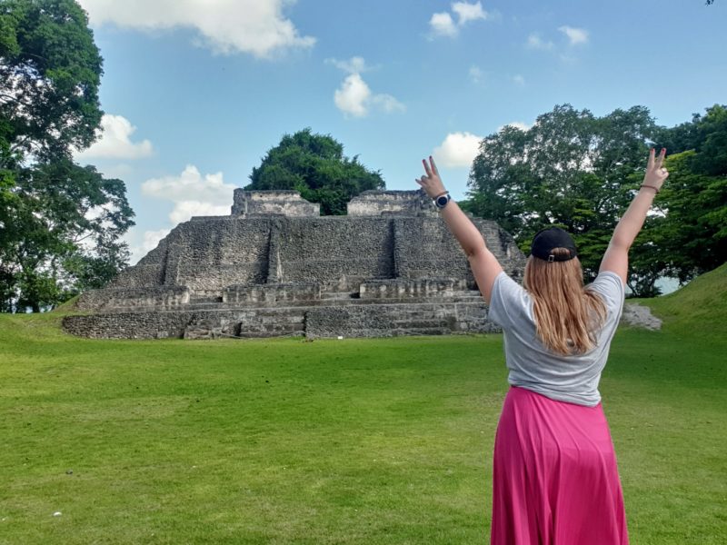Roma wearing a fuscia pink midi skirt and a grey tshirt and a black cap standing in front of Maya ruins Xunantunich ruins in Belize