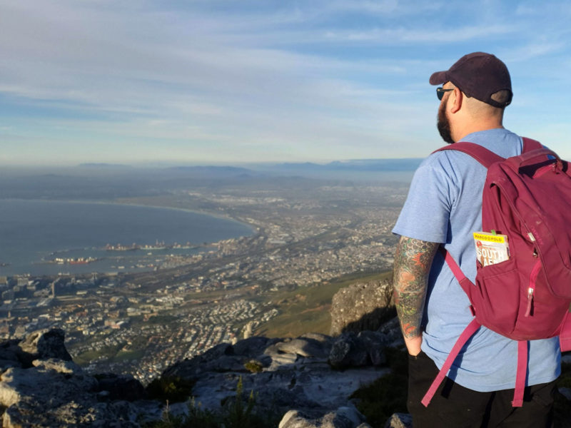 Russ viewing Cape Town from the top of Table Mountain in Cape Town. He's wearing a light blue shirt and wearing a purple backpack