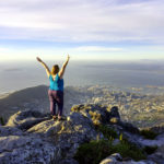 Roma standing on the edge of Table Mountain in Cape Town with her arms in the arm in celebration