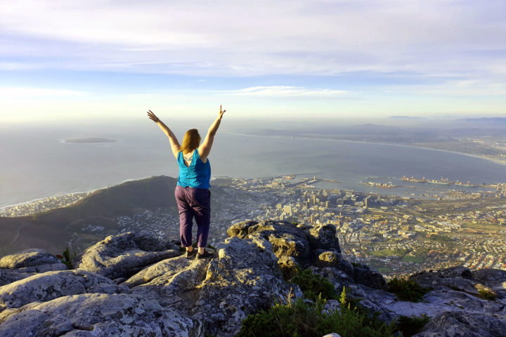 Roma standing on the edge of Table Mountain in Cape Town with her arms in the arm in celebration