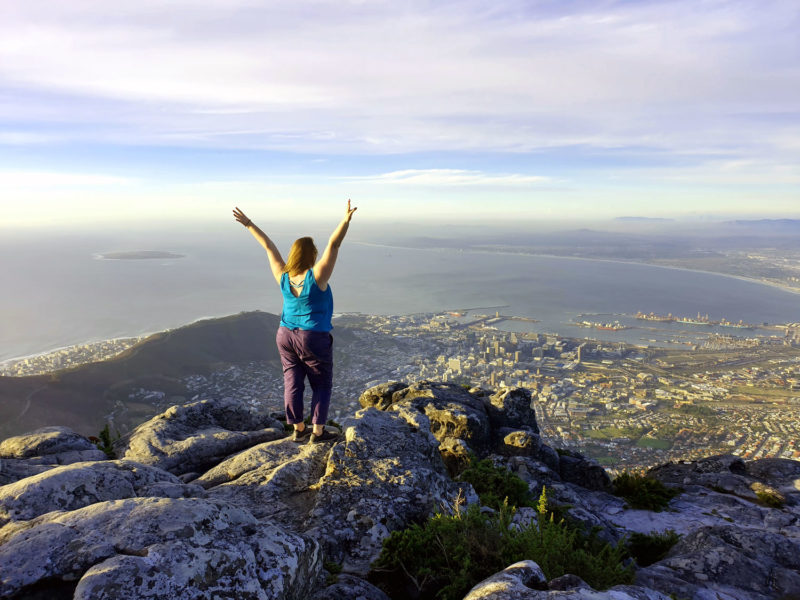 Roma standing on the edge of Table Mountain in Cape Town with her arms in the arm in celebration