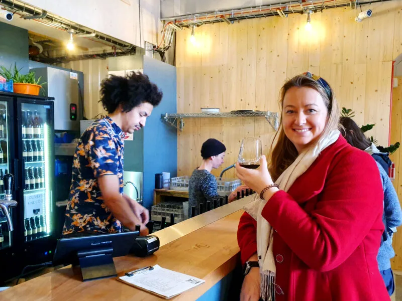 Roma standing at the bar in a red jacket holding a beer at the Kernel on the Bermondsey Beer Mile