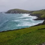 Visit the Dingle Peninsula. View of coastline with light blue waves on the left with dark green grass-covered cliffs on the right