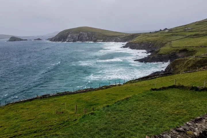Visit the Dingle Peninsula. View of coastline with light blue waves on the left with dark green grass-covered cliffs on the right