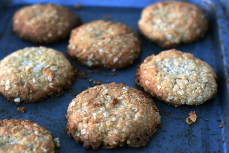 Close up of ANZAC cookie on dark tray which shows two biscuits in focus and four out of focus. All created with a chewy ANZAC biscuit recipe.