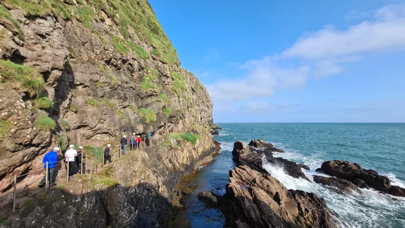 Line of people walking along The Gobbins cliff path with sea views and blue skies. 