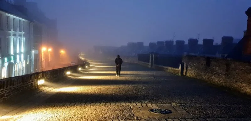 Russell walking away from the camera along the city walls of Derry at dusk. The atmosphere is foggy and spooky.